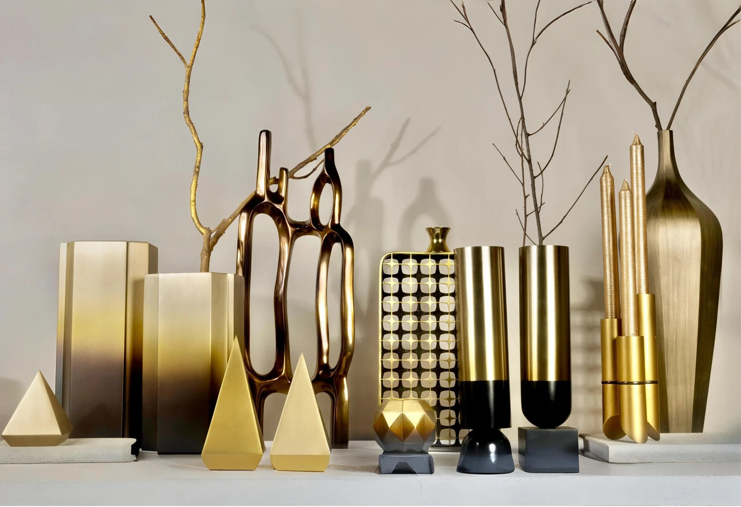 Luxury modern home decor accessories including brushed gold vases, brass cylindrical containers, geometric pyramid sculptures, and metallic decorative objects for contemporary interior design